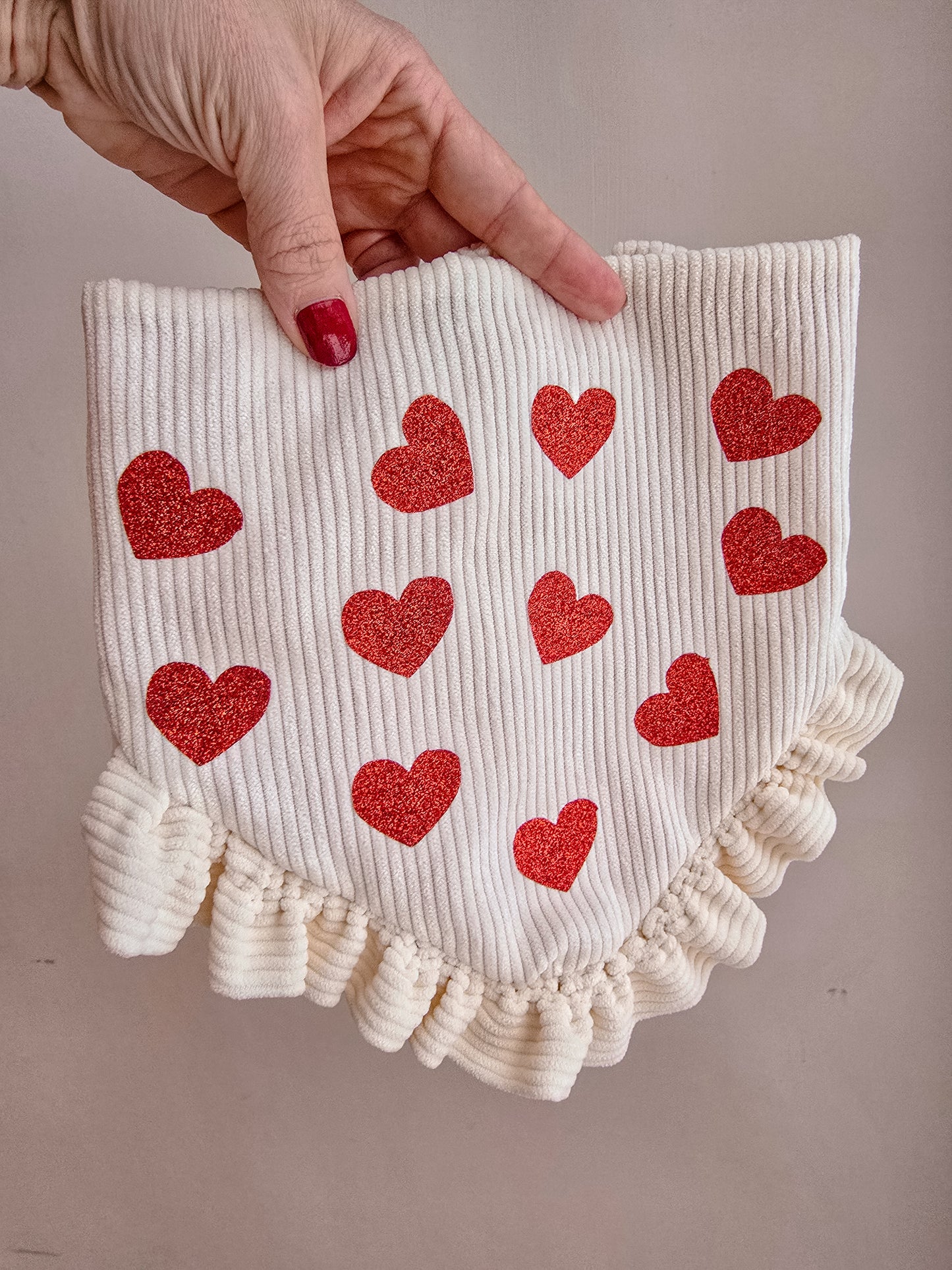Dog wearing a cream corduroy scrunchie bandana with glittery red hearts and ruffles, surrounded by Valentine's Day-themed decor.
Alt Text for Image 2 (Reverse Side):
Hand holding a reversible scrunchie dog bandana with cream corduroy and red glitter hearts on one side, and a red fabric with white polka-dot hearts on the reverse, featuring delicate ruffles and a branded logo tag.
