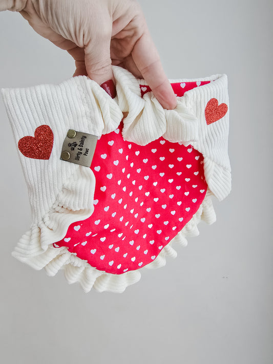 Dog wearing a cream corduroy scrunchie bandana with glittery red hearts and ruffles, surrounded by Valentine's Day-themed decor.
Alt Text for Image 2 (Reverse Side):
Hand holding a reversible scrunchie dog bandana with cream corduroy and red glitter hearts on one side, and a red fabric with white polka-dot hearts on the reverse, featuring delicate ruffles and a branded logo tag.