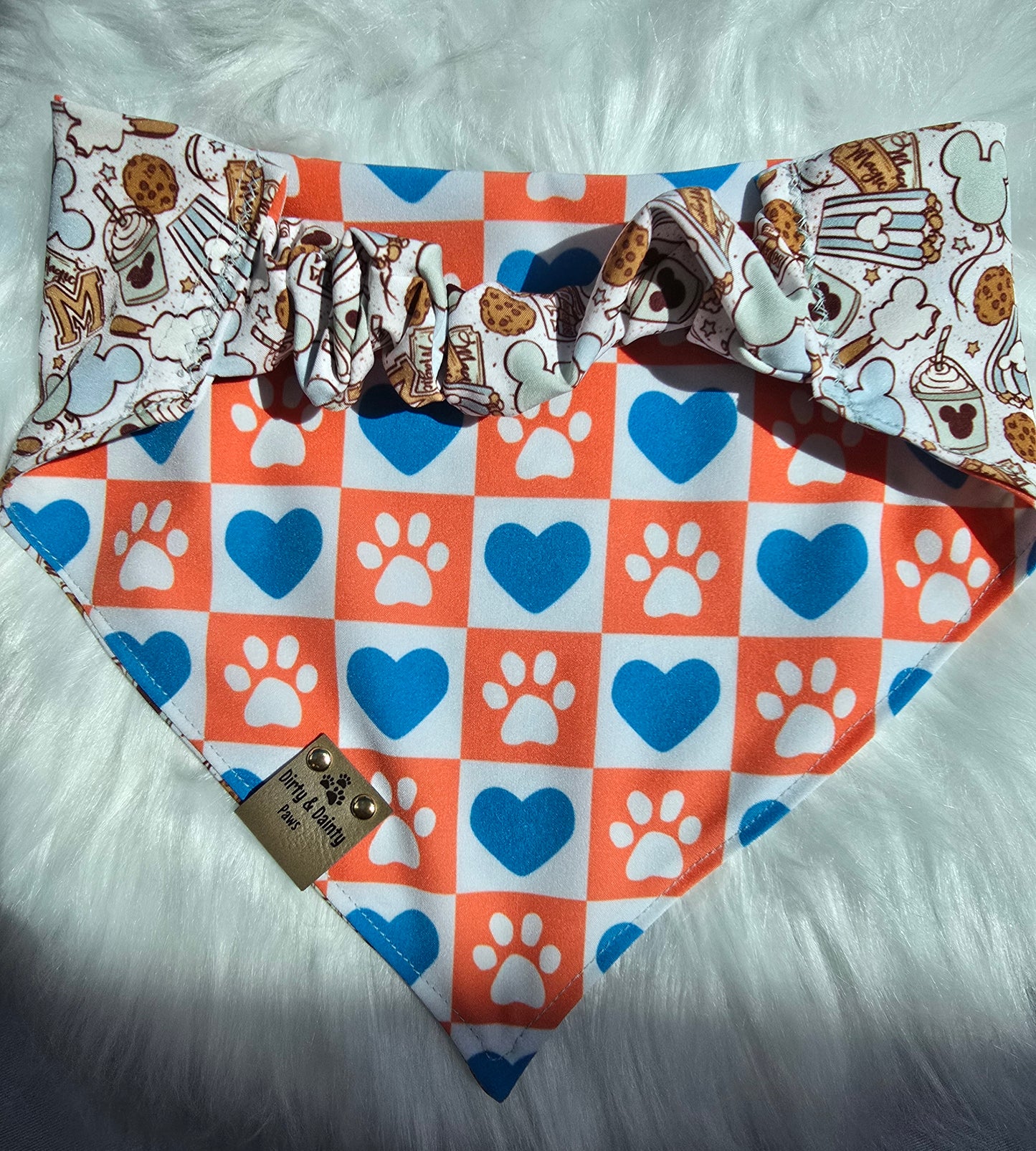 Close-up of Magical Treats Disney-inspired with Reversible dide blue and orange checkers with hearts and paw prints dog bandana with faux leather Dirty & Dainty Paws tag, made with Zoomie Proof™ polyester fabric.