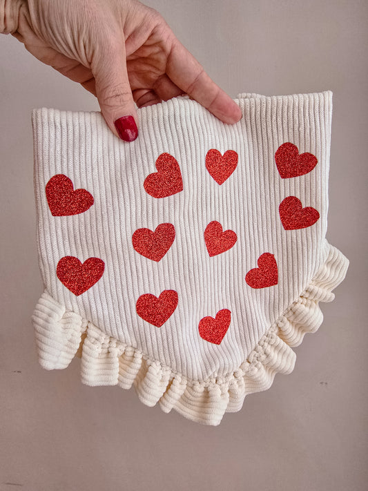 Dog wearing a cream corduroy scrunchie bandana with glittery red hearts and ruffles, surrounded by Valentine's Day-themed decor.

Alt Text for Image 2 (Reverse Side):
Hand holding a reversible scrunchie dog bandana with cream corduroy and red glitter hearts on one side, and a red fabric with white polka-dot hearts on the reverse, featuring delicate ruffles and a branded logo tag.