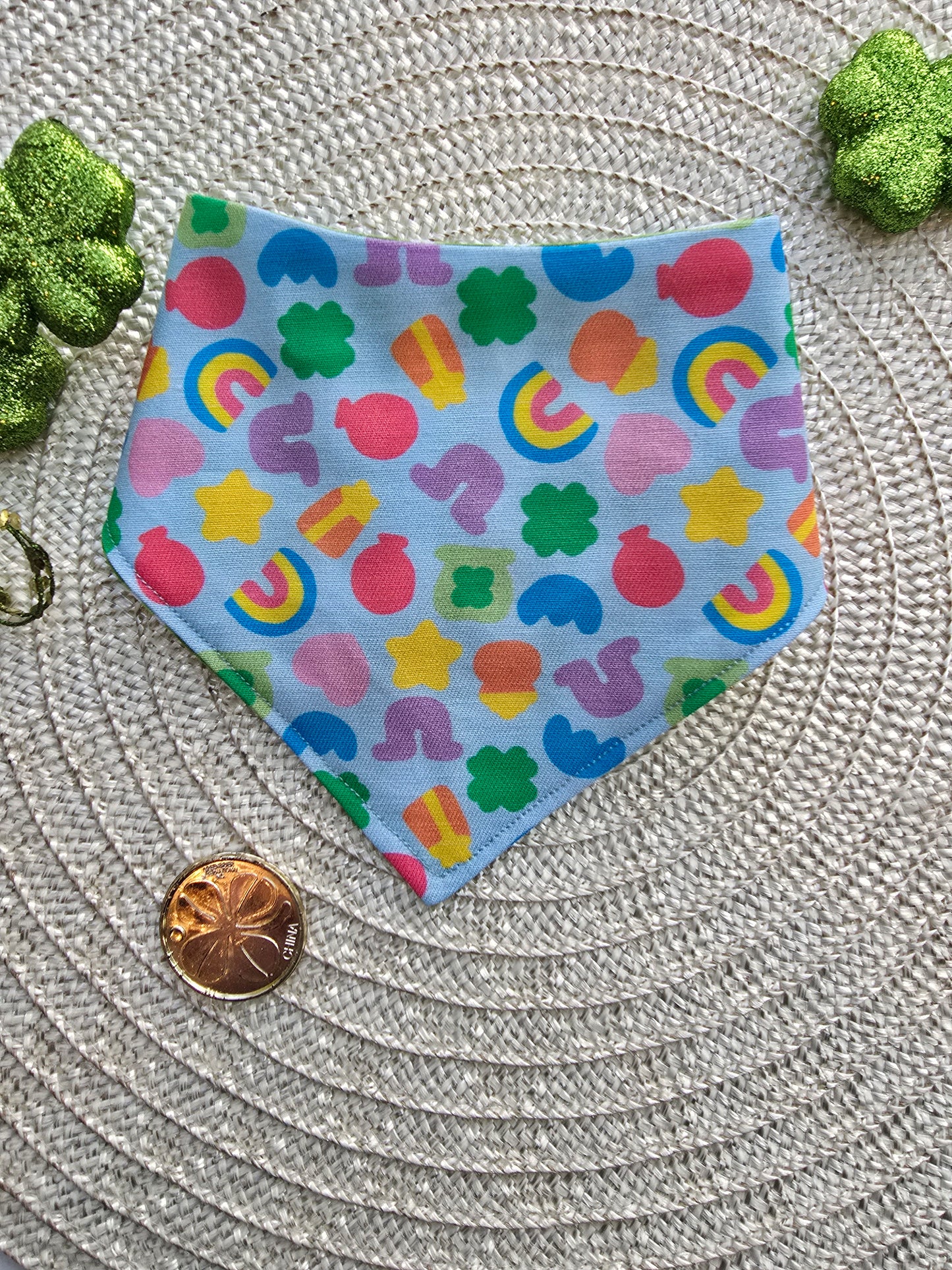 A colorful scrunchie-style dog bandana with a Lucky Charms-inspired pattern, featuring rainbows, stars, clovers, and horseshoes on a light blue background, laid flat on a woven mat with St. Patrick’s Day decorations