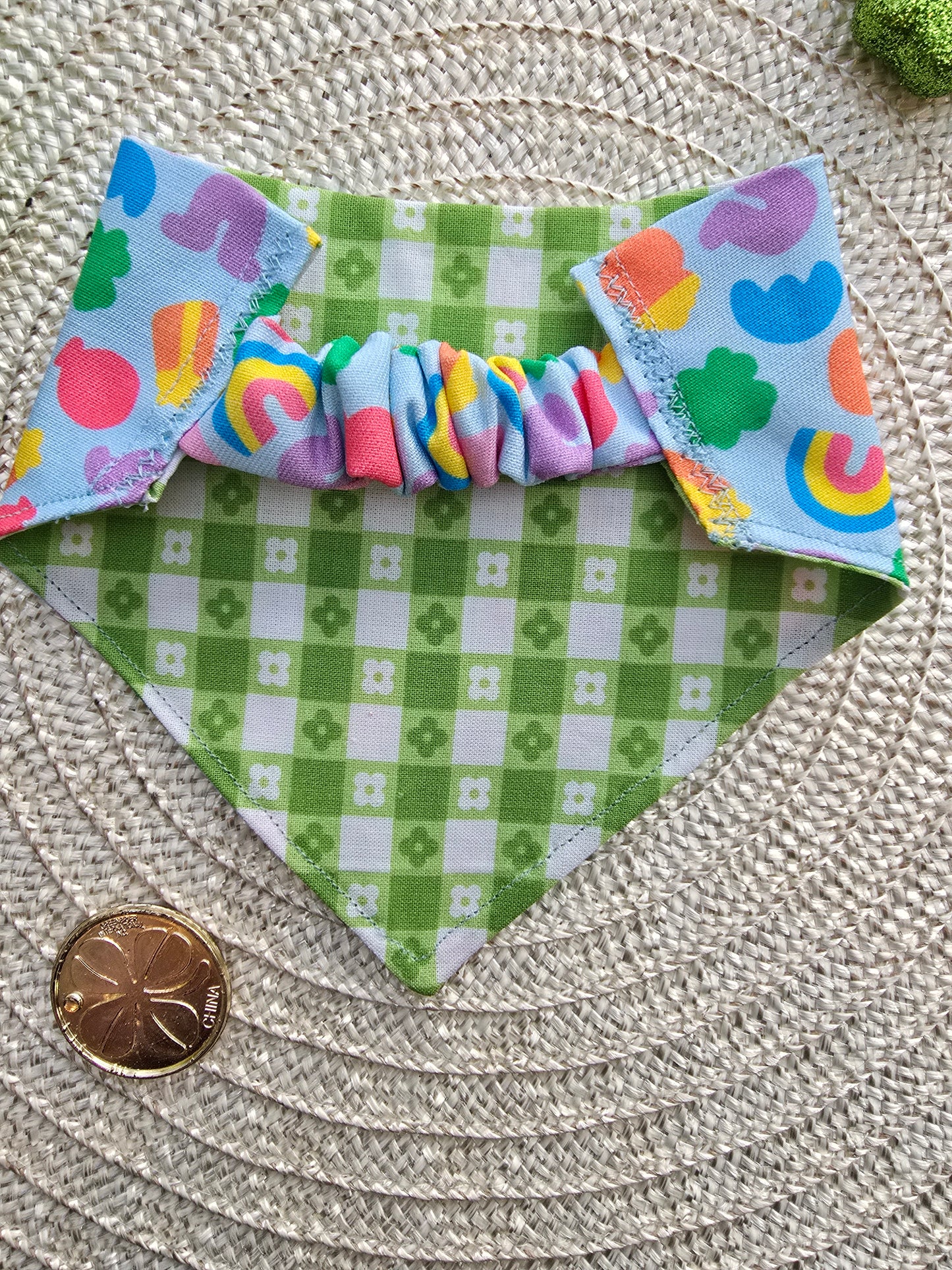 The reversible side of the scrunchie dog bandana, showcasing a green and white gingham clover pattern. The stretchy scrunchie-style opening is visible, allowing for easy slip-on wear. Displayed on a woven mat with festive St. Patrick’s Day decor.