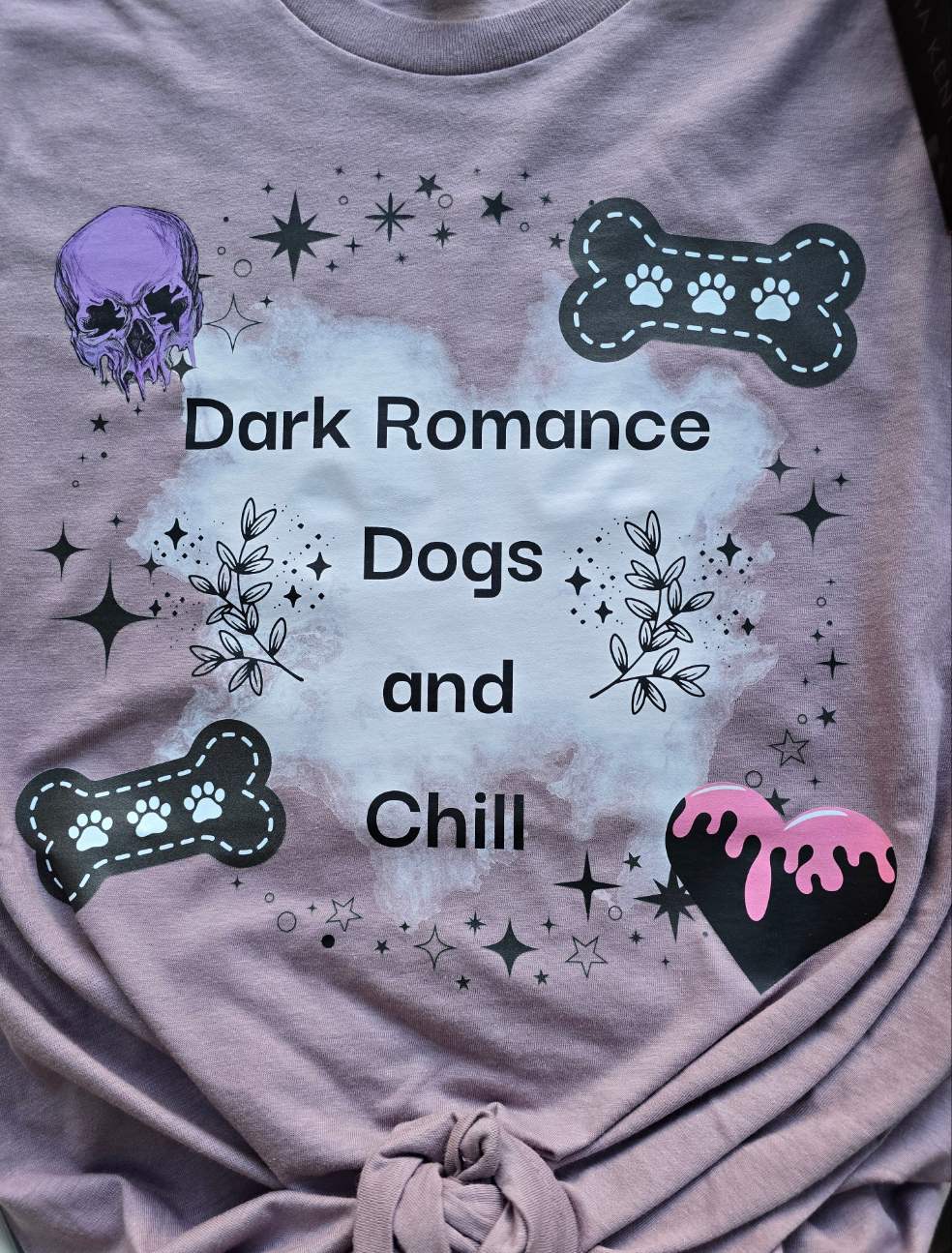 A soft mauve-colored t-shirt with the words "Dark Romance Dogs and Chill", featuring gothic-inspired designs like a skull, bones, and stars. Styled with dark romance books.