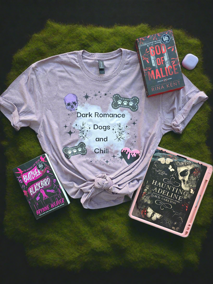 A soft mauve-colored t-shirt with the words "Dark Romance Dogs and Chill", featuring gothic-inspired designs like a skull, bones, and stars. Styled with dark romance books.