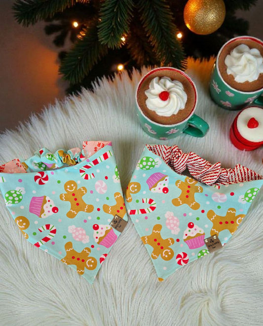 Two colorful dog bandanas with gingerbread and candy cane patterns on a fluffy white surface with hot chocolate and cookies.