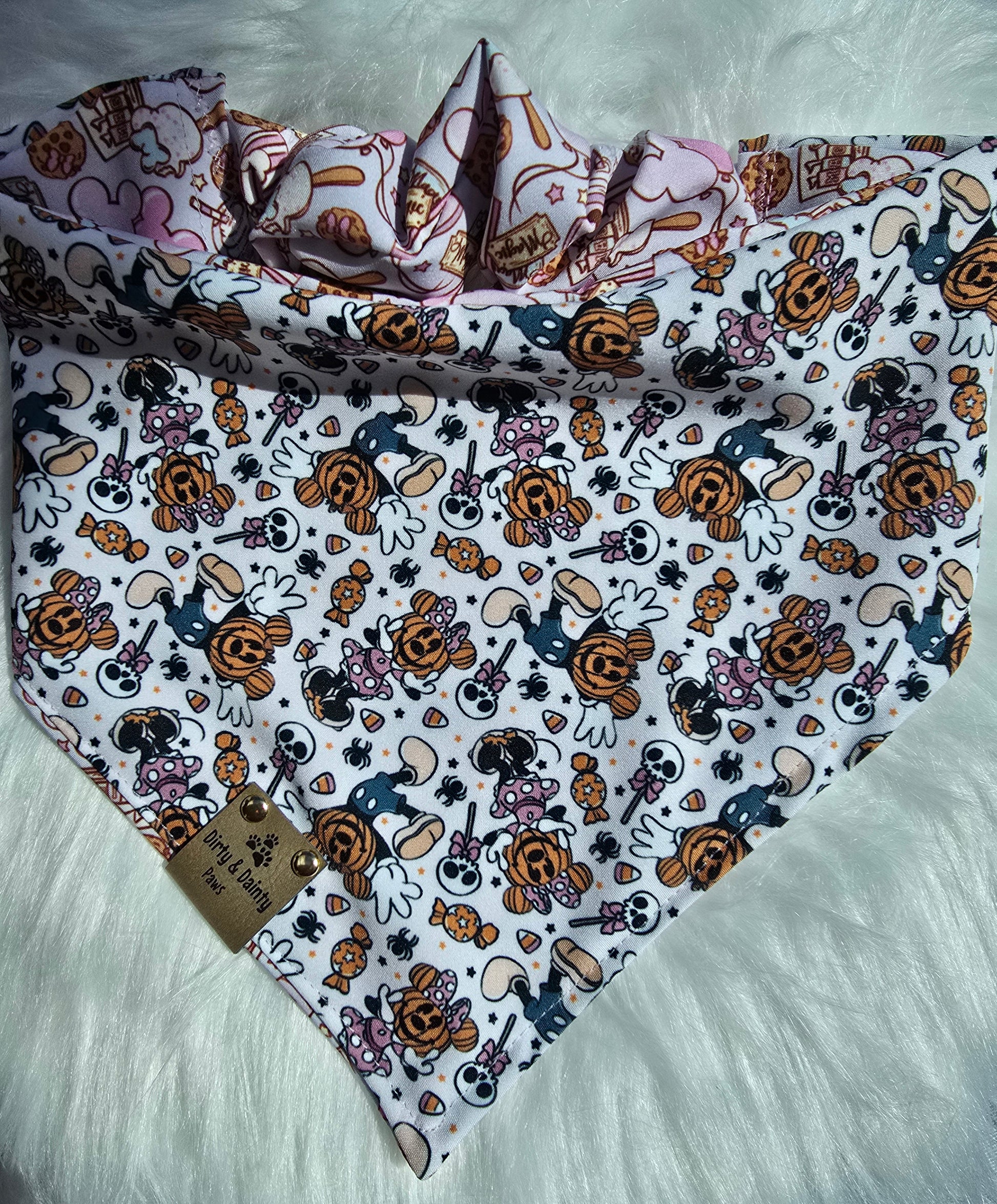 Close-up of Magical Treats Disney-inspired mickey and Minnie jack-o-lantern  dog bandana with faux leather Dirty & Dainty Paws tag, made with Zoomie Proof™ polyester fabric.