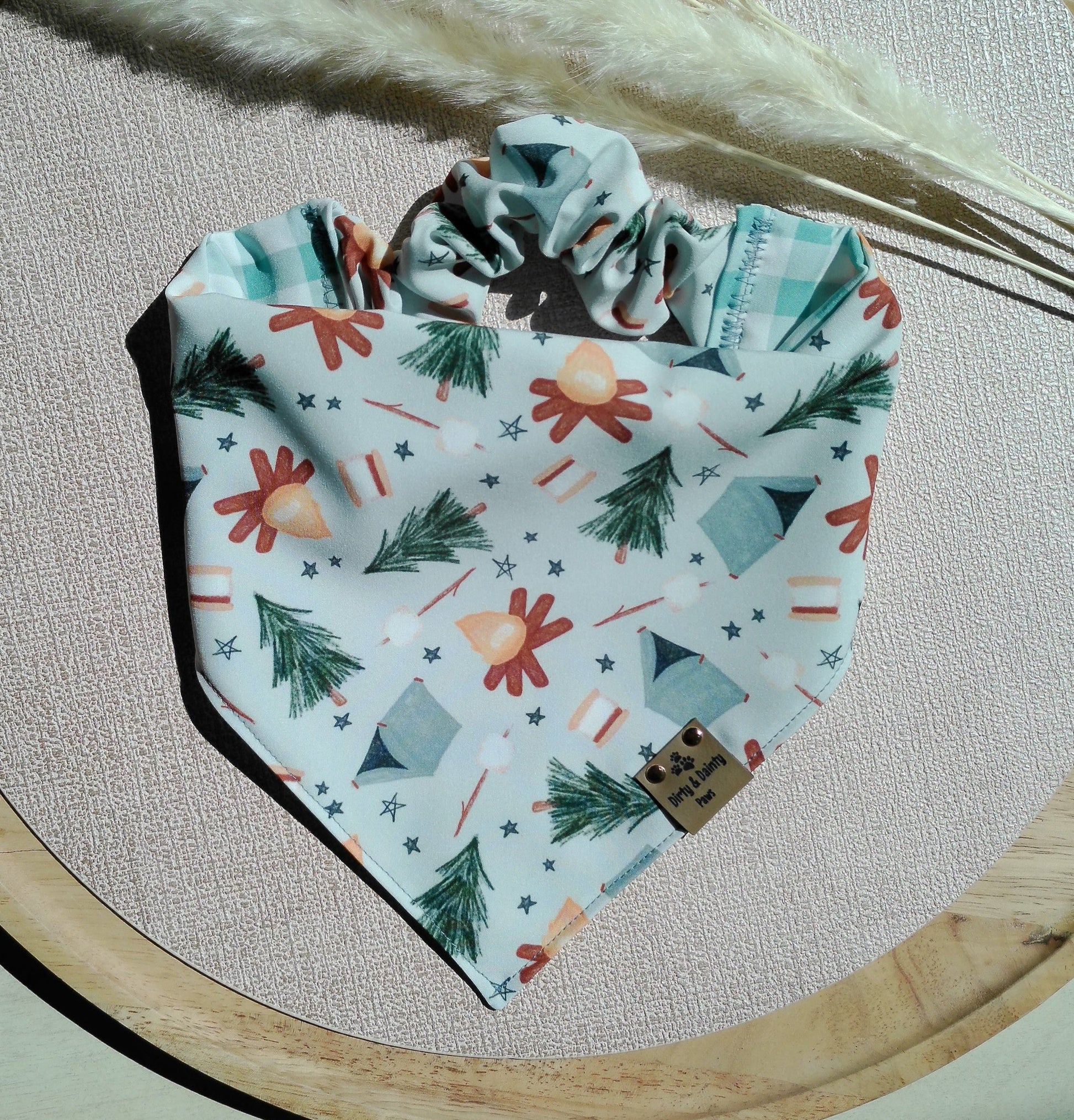 Reversible ZoomieProof™ dog bandana featuring camping icons including tents, campfires, lanterns, pine trees, and marshmallows with a coordinating mint gingham reverse side. Handmade scrunchie dog bandana by Dirty and Dainty Paws