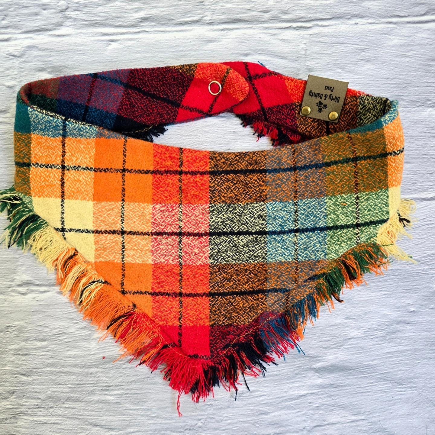 Colorful plaid dog bandana made from high-quality flannel, featuring hand-fringed edges, a secure snap closure, and a faux leather logo tag. Perfect for fall, the bandana showcases a vibrant mix of autumnal colors.